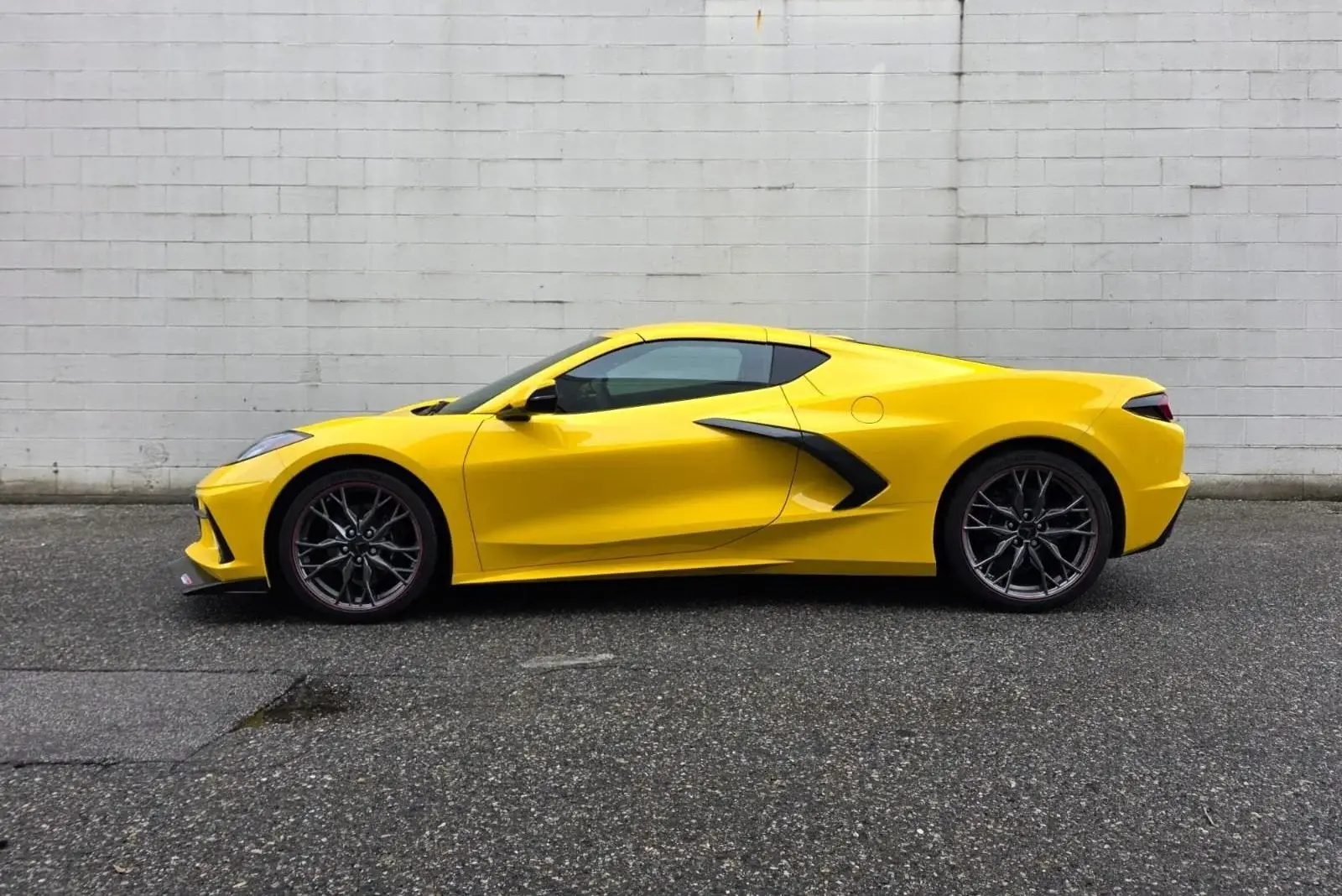 Side profile of a yellow Chevrolet Corvette C8 with gloss paint, parked against a white block wall
