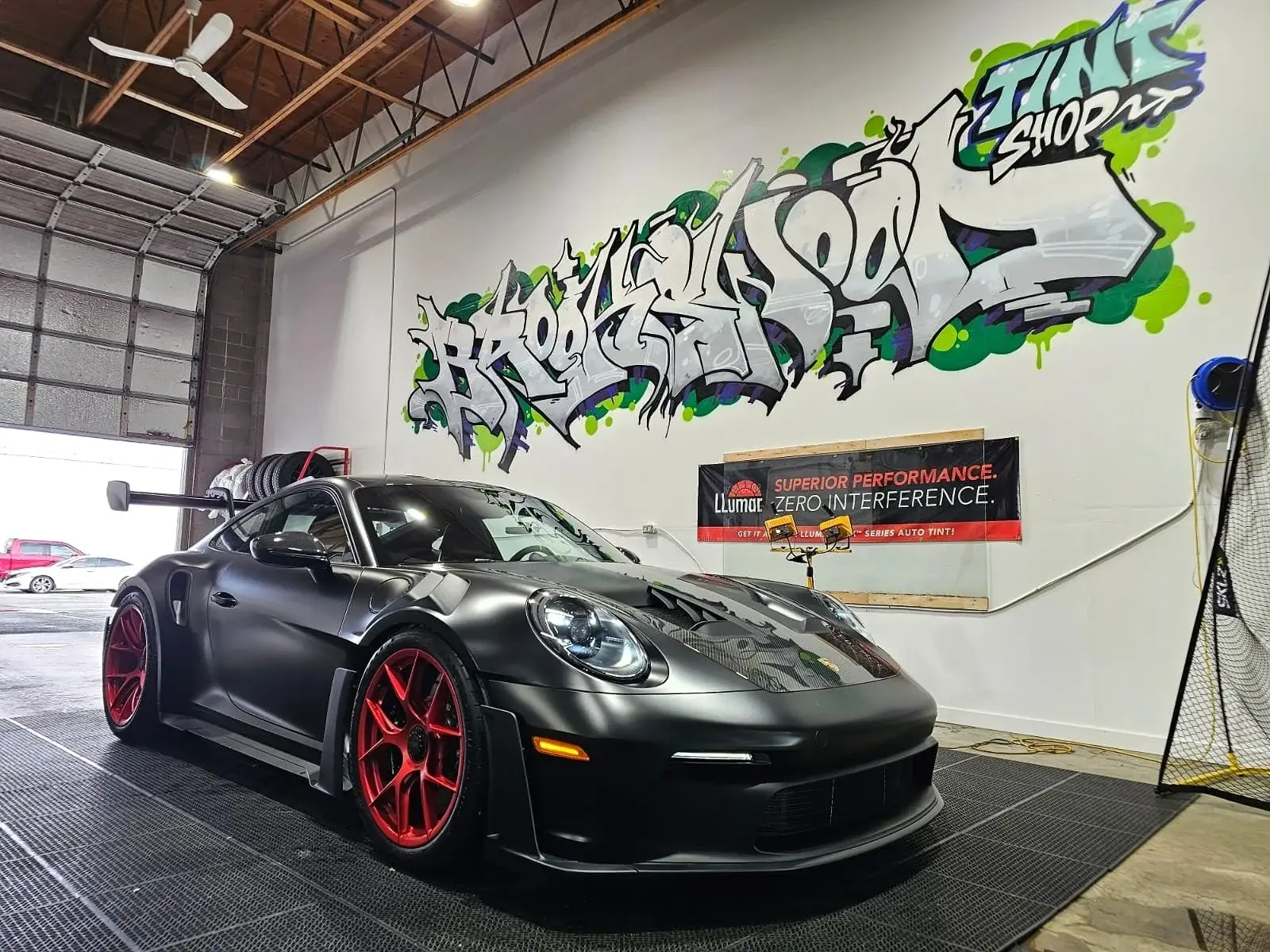 Matte black Porsche 911 GT3 RS with red wheels inside Brookswood Tint Shop, graffiti wall behind