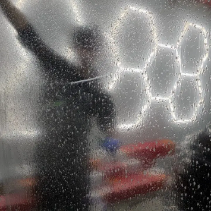 An installer seen as a blurred silhouette through a sheet of wet window film, hexagonal shop lights glowing behind him