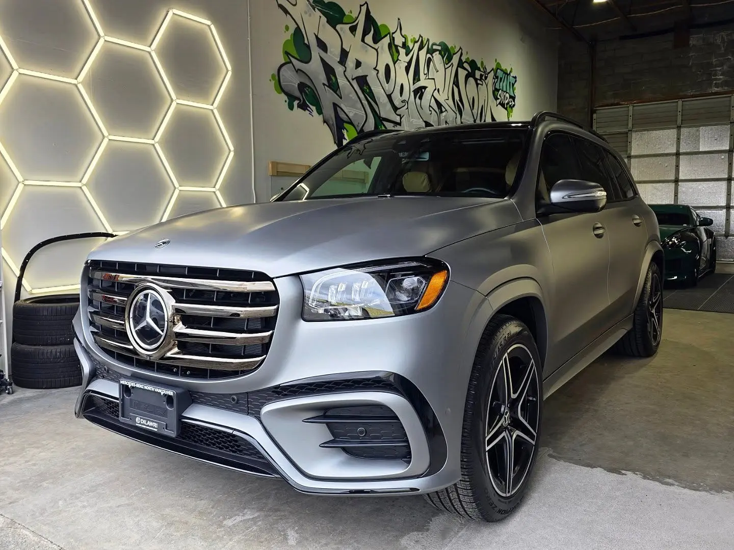 Matte grey wrapped Mercedes GLS parked in the shop's enclosed bay under hexagon lights, with a roll-up door shut to the outside
