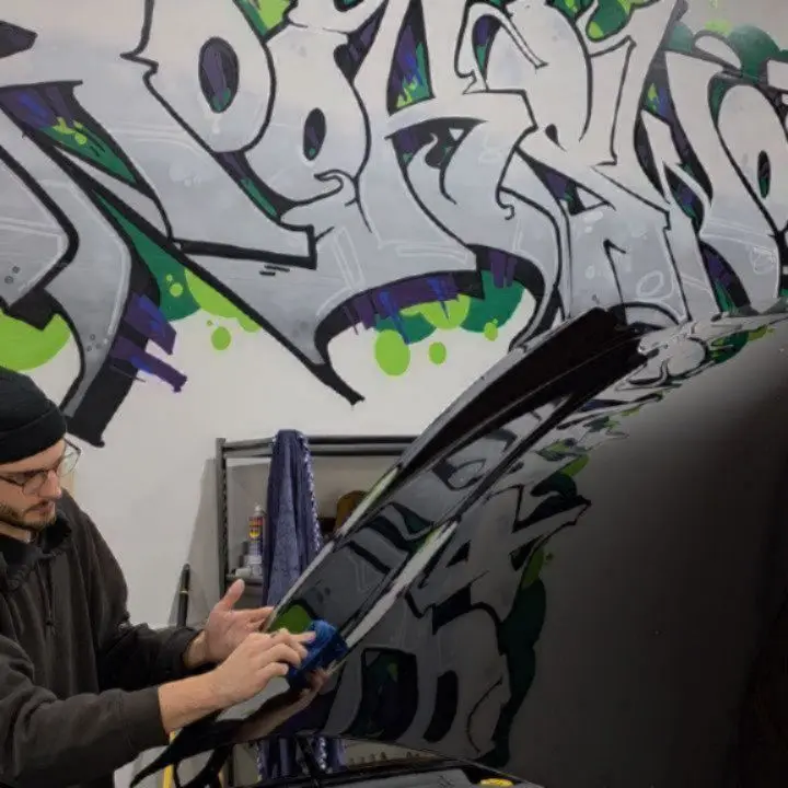 Technician in a beanie and glasses wiping the black hood of a car with a blue microfibre towel under the shop's graffiti mural in Langley