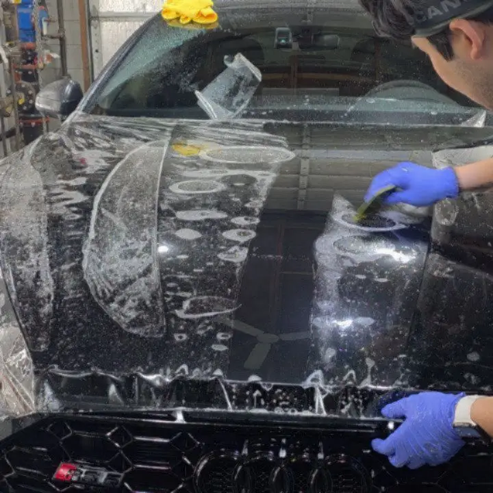 Gloved hands squeegeeing paint protection film onto a black hood