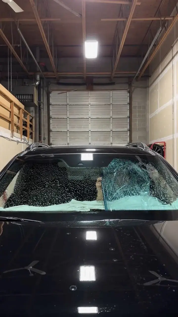 Black SUV in the shop with film being smoothed onto the inside of its windshield, a hand visible through the wet glass and towels on the dash