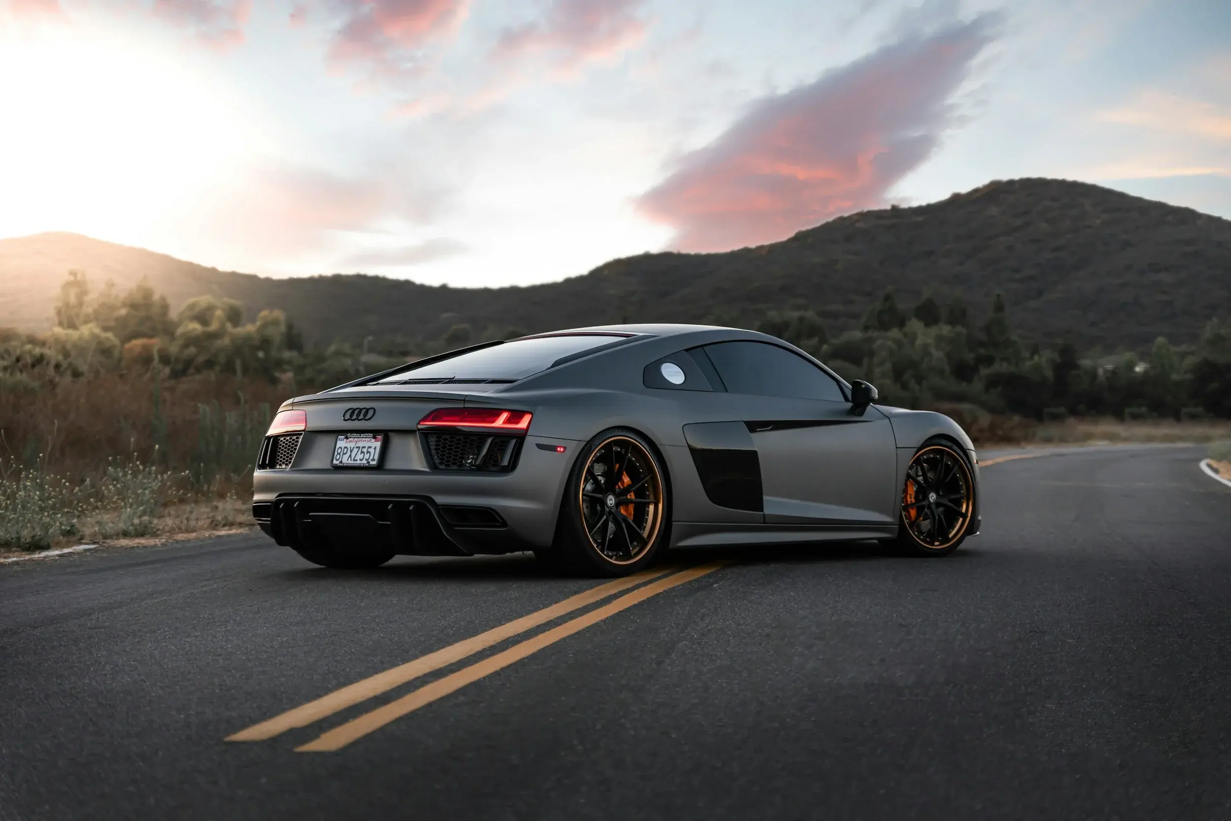 Flat grey Audi R8 coupe on a hillside road at sunset, seen from the rear three-quarter