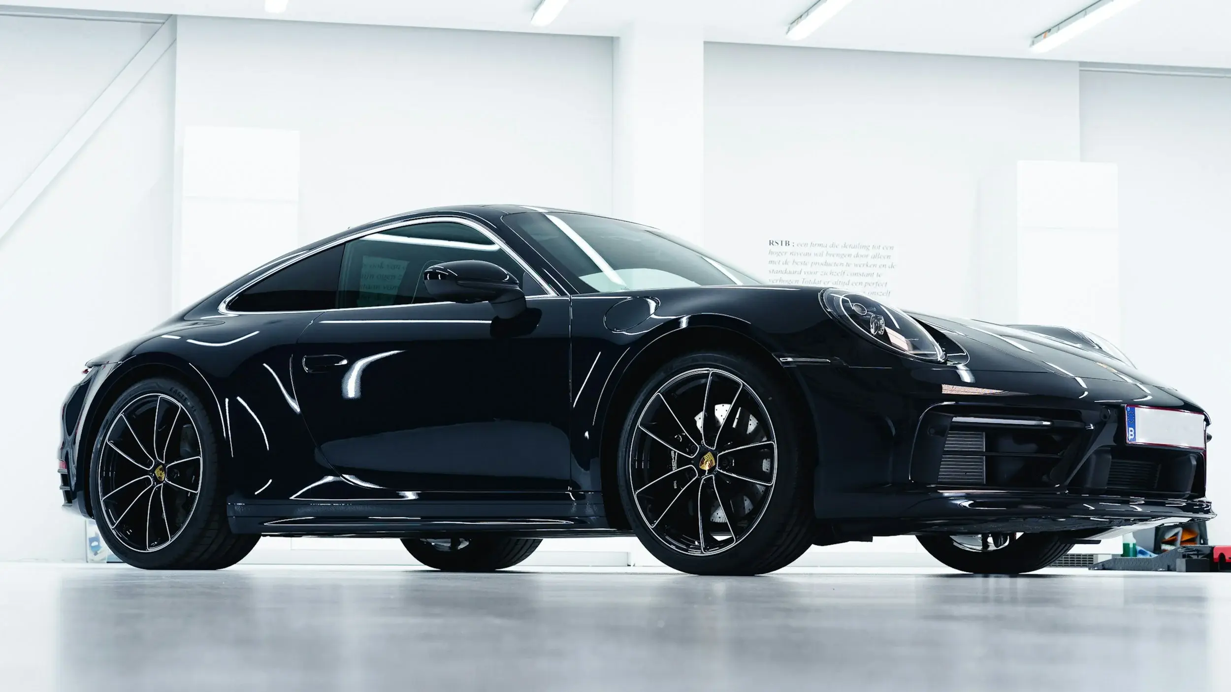 Black Porsche 911 coupe in a bright white studio, overhead lights reflecting off its gloss doors and front fender