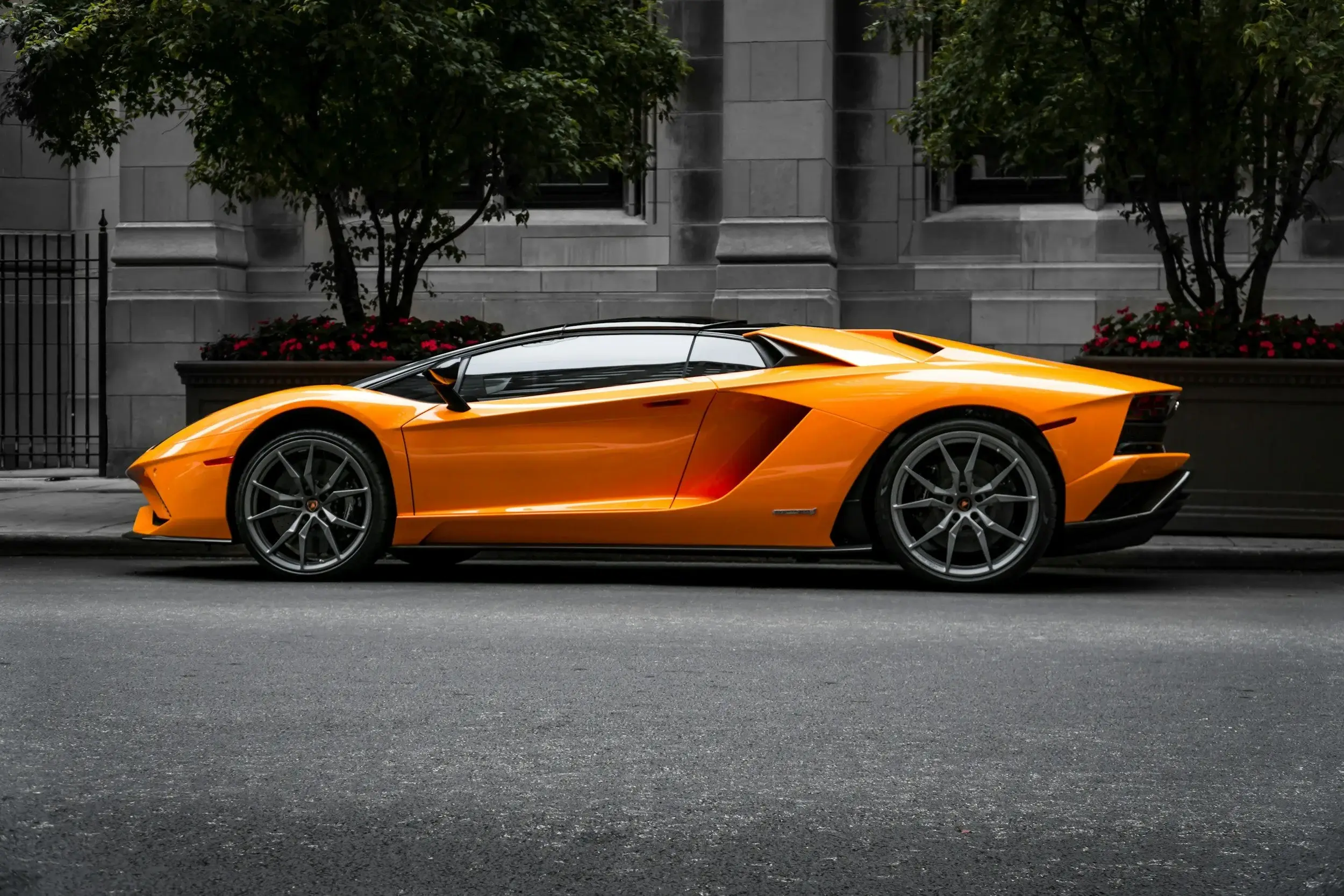 Orange Lamborghini Aventador parked at a city curb beside planters and street trees