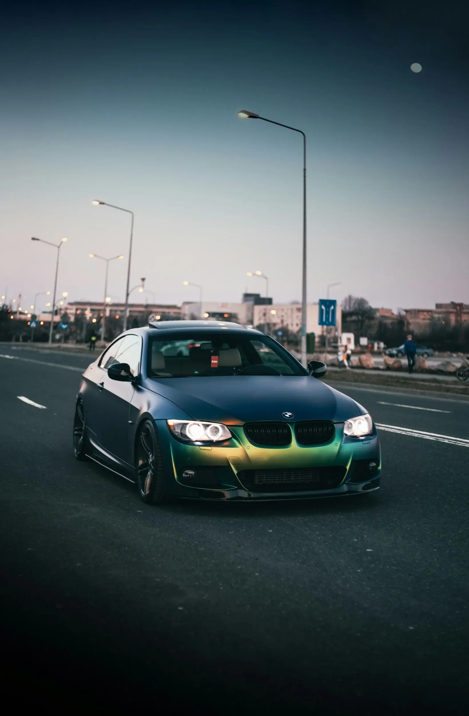 BMW coupe in a green-to-blue colour-shift finish on a city street at dusk, headlights on