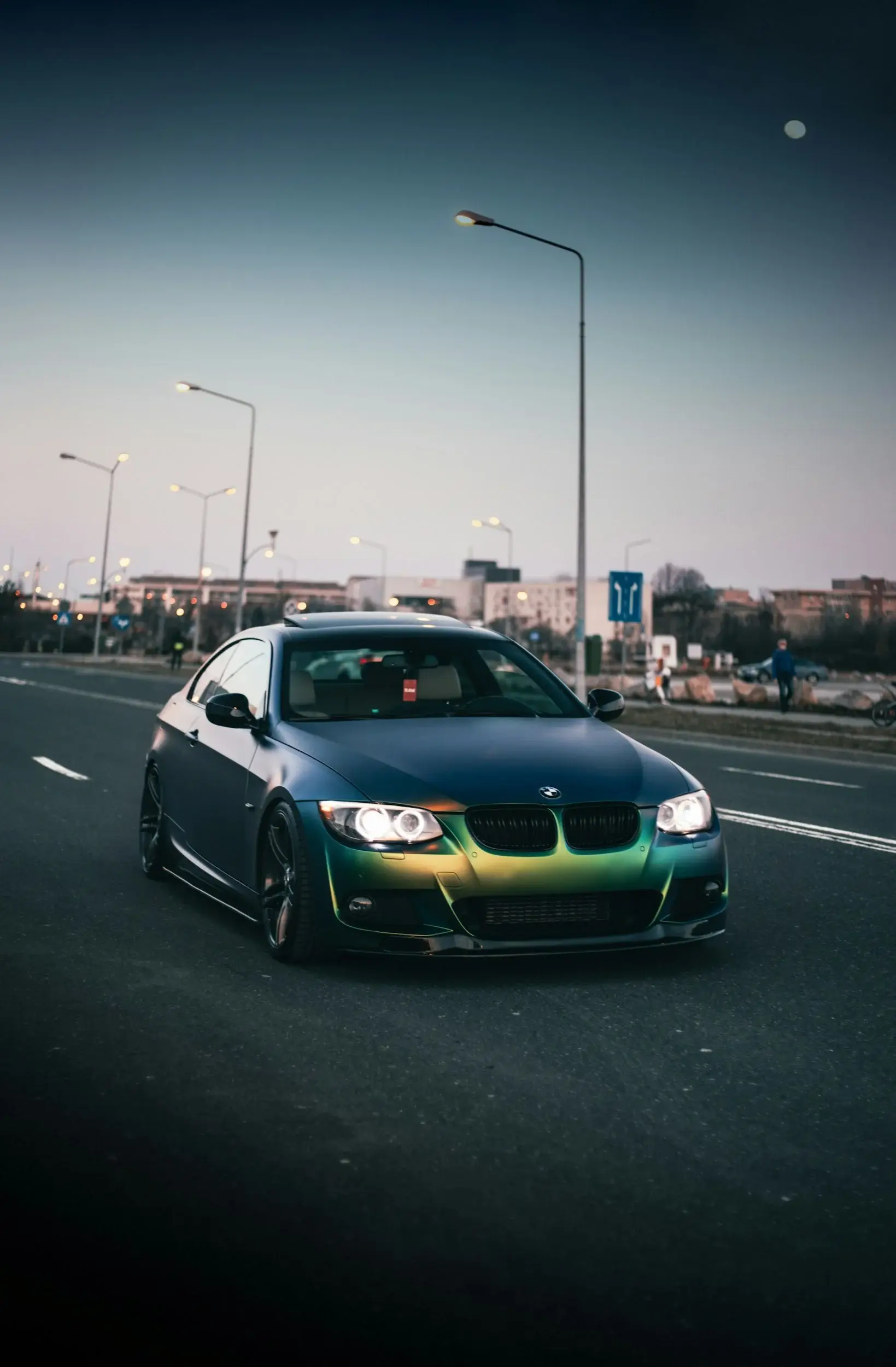 Colour-shift wrapped BMW coupe with its headlights on, on a city road at dusk under lit street lamps