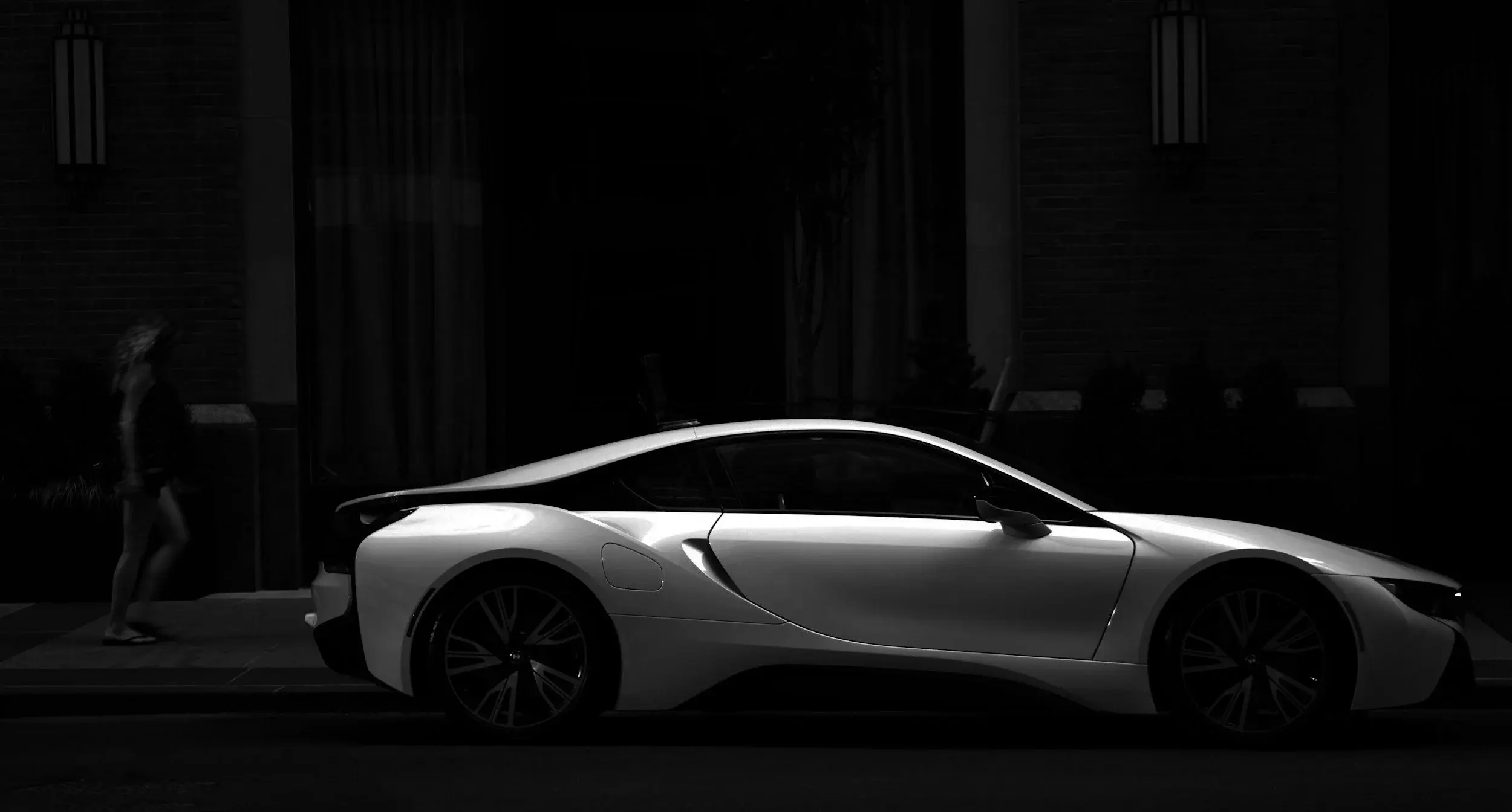 Black and white side view of a BMW i8 plug-in hybrid coupe parked on a dark city street as a pedestrian walks past