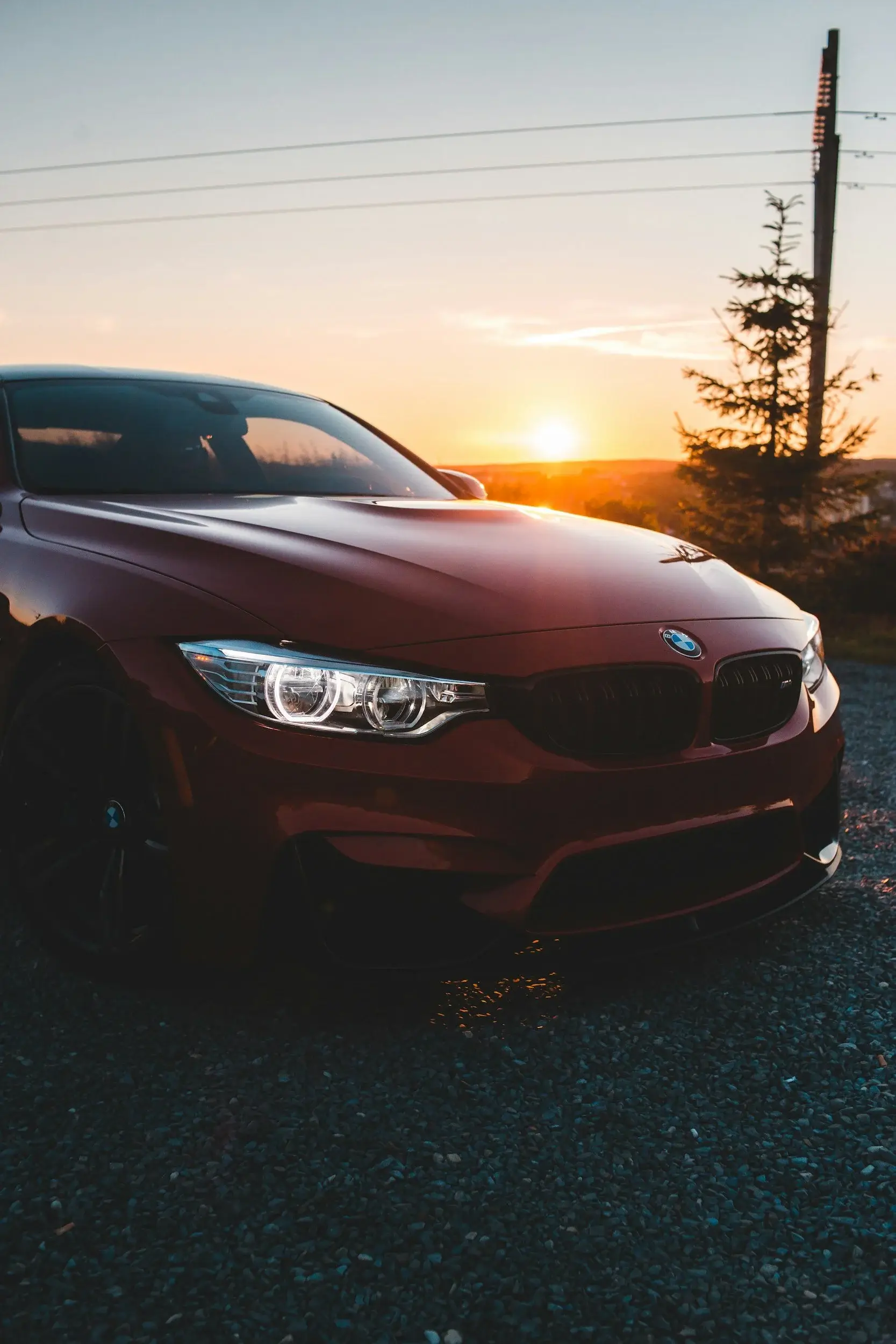 Front bumper, hood and headlight of a red BMW M4 parked on loose gravel at sunset