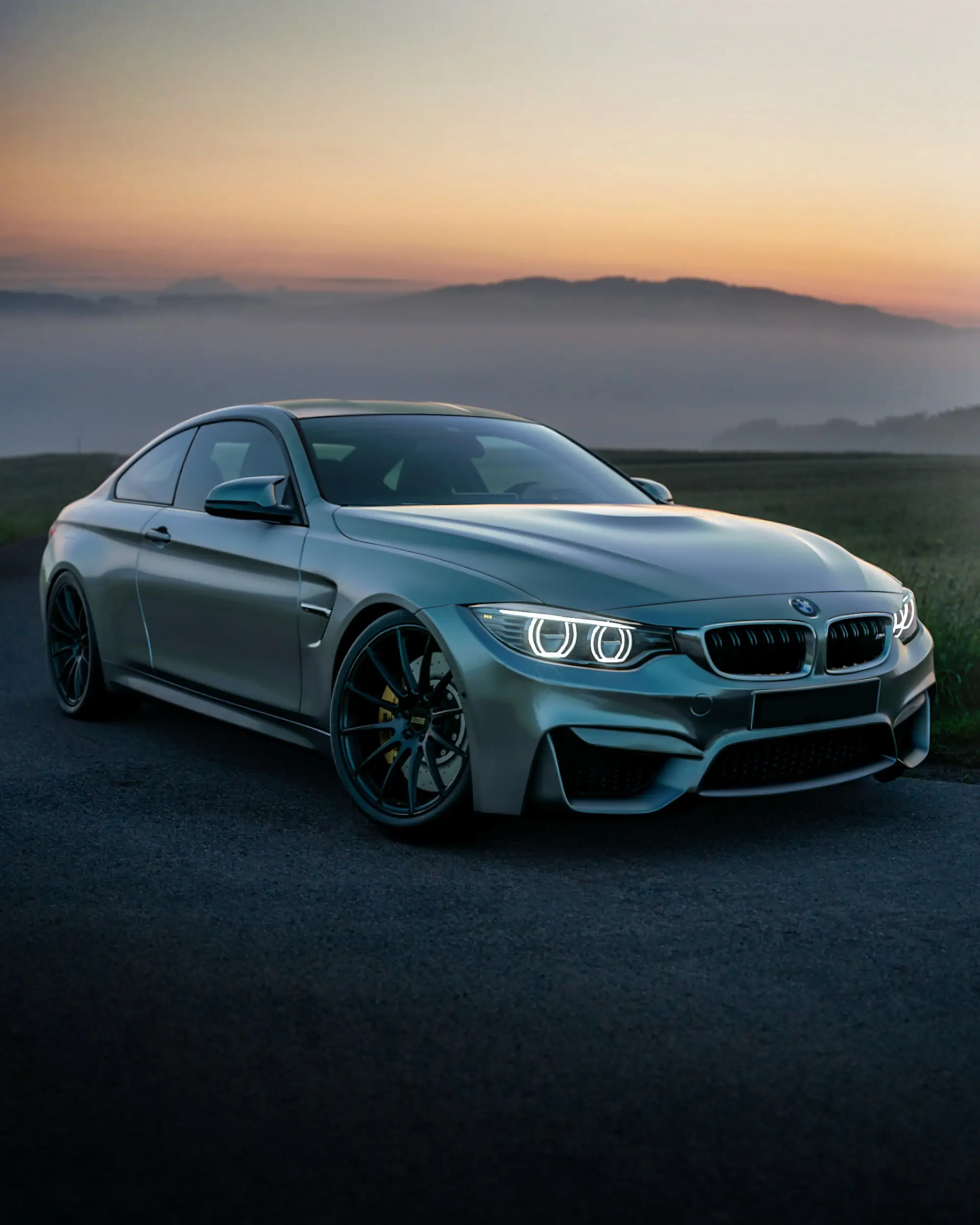Grey BMW M4 coupe parked on a country road at sunset, low light across its hood and front bumper