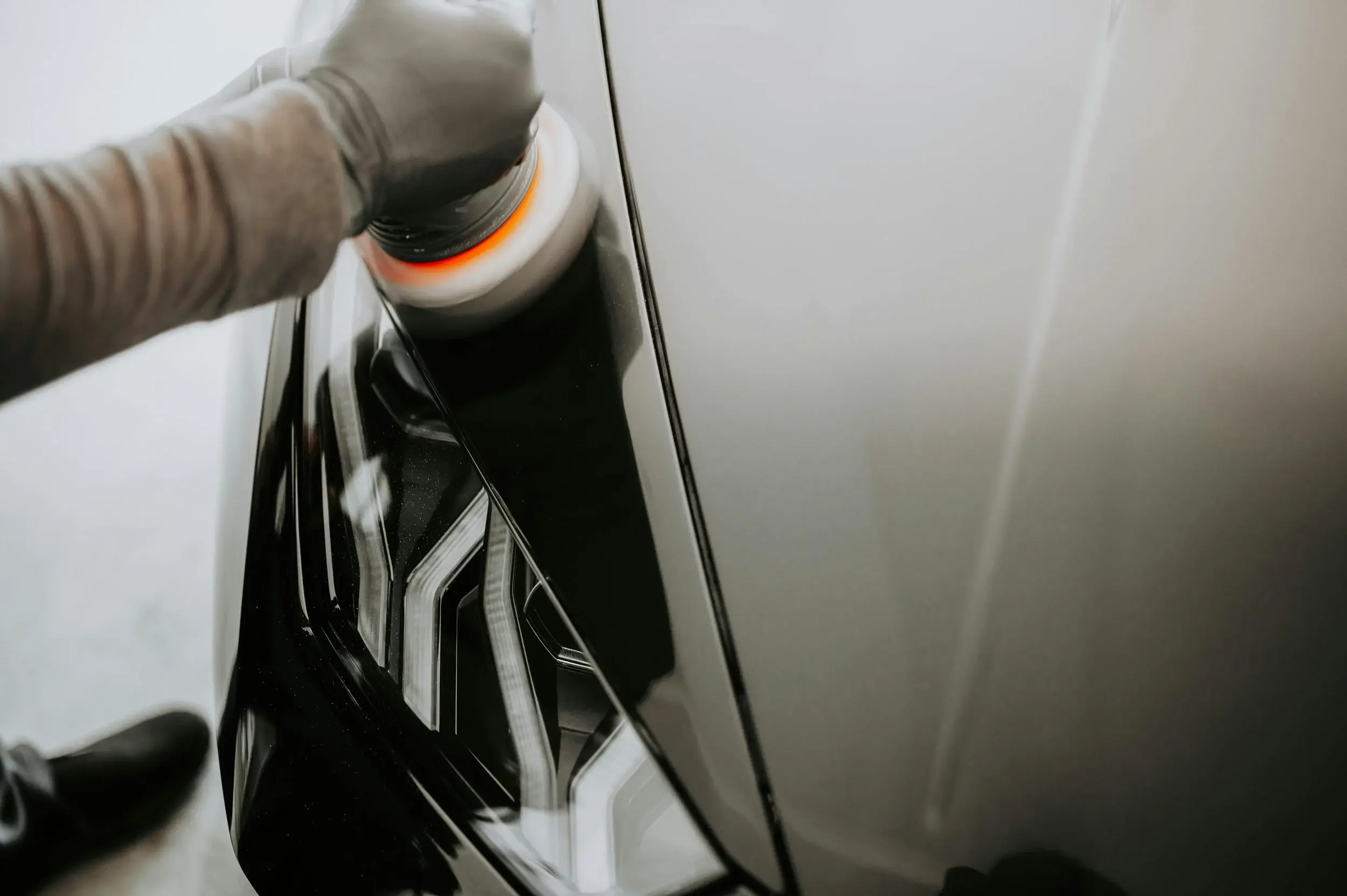 Gloved hand holding an orbital polisher against a light grey car next to its black lamp housing