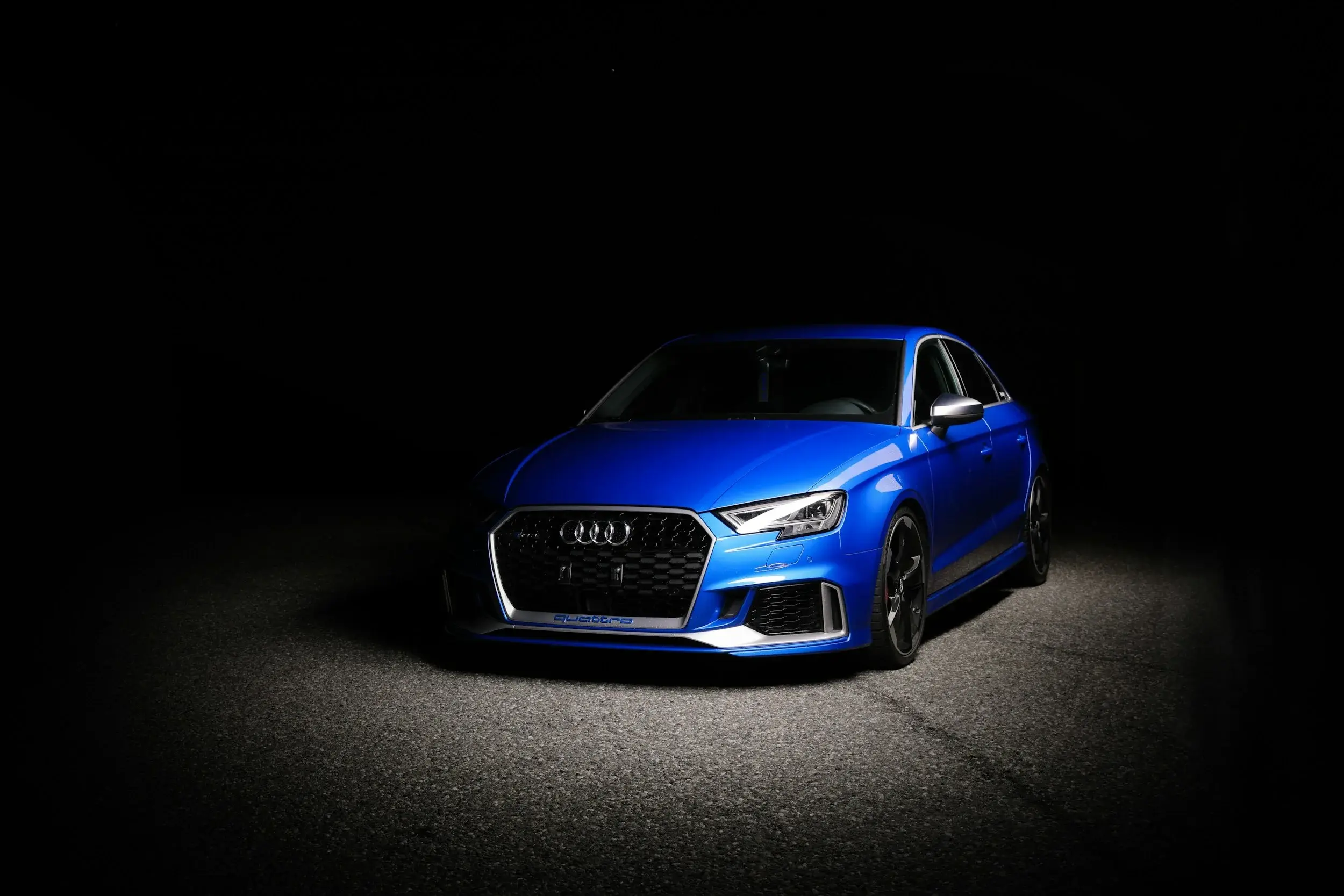 Blue Audi RS 3 sedan in a dark studio, light falling across its glossy hood and front bumper