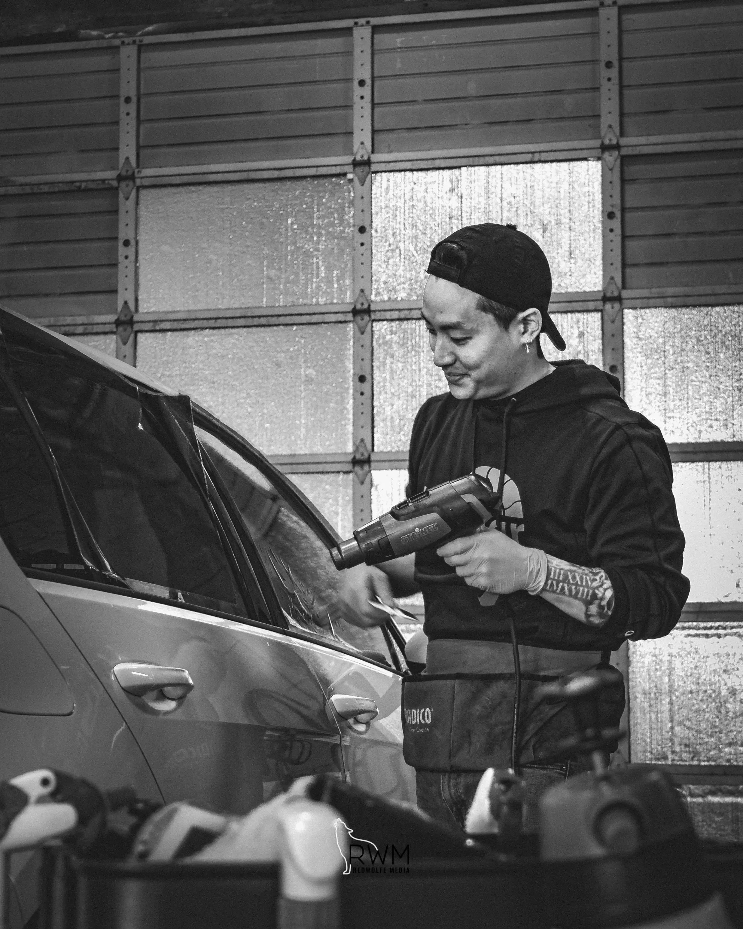 Smiling technician with a heat gun beside a car in the shop, black and white